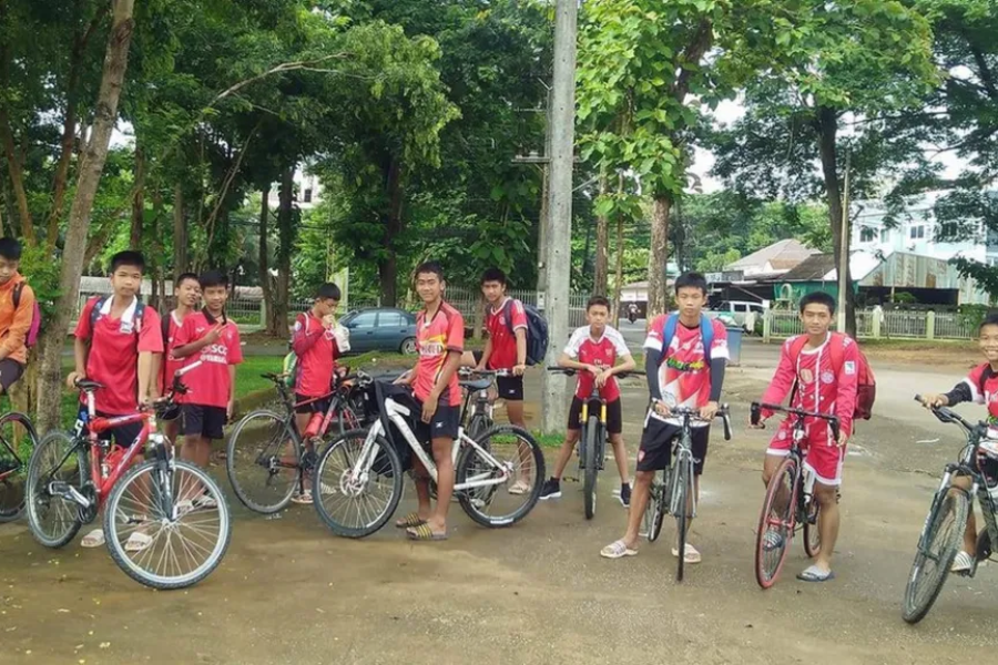 A group of boys wearing red sports jerseys pose with their bicycles in a shaded outdoor area surrounded by trees. Most of them wear backpacks and flip-flops, and they appear to be part of a team or school group, gathered on a dirt clearing near a quiet road and parked cars.
