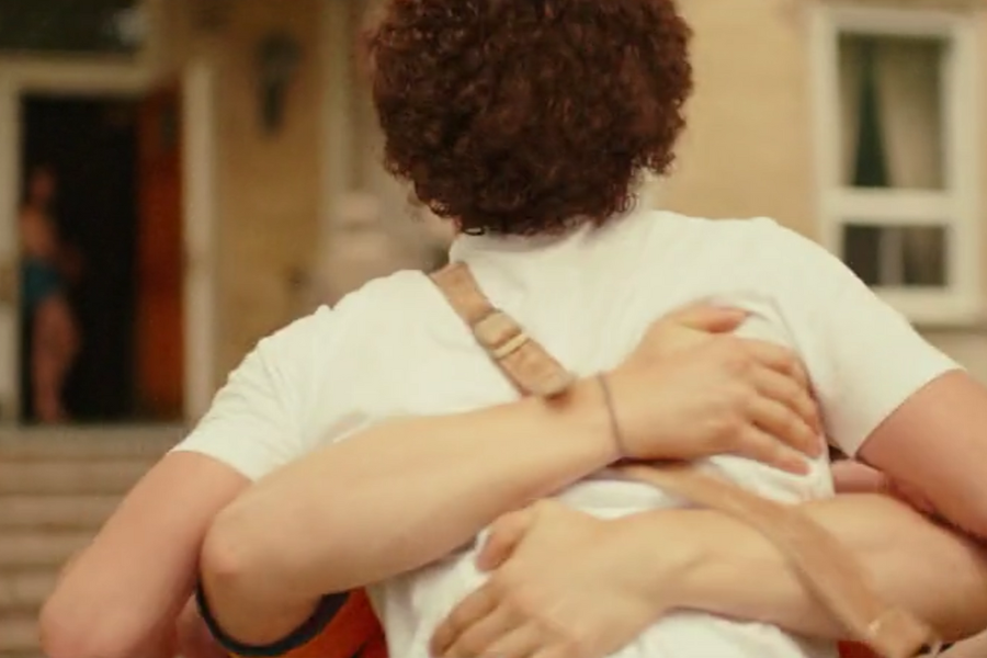 Two people tightly embrace outside a building, with one wearing a white t-shirt and tan suspenders. The warm, close hug is framed from behind, capturing a moment of deep connection and affection.