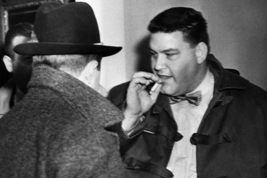 Black and white photo of a man in a bow tie and heavy coat mid-conversation, holding a cigarette near his mouth. Another man in a wide-brimmed hat and overcoat has his back to the camera.