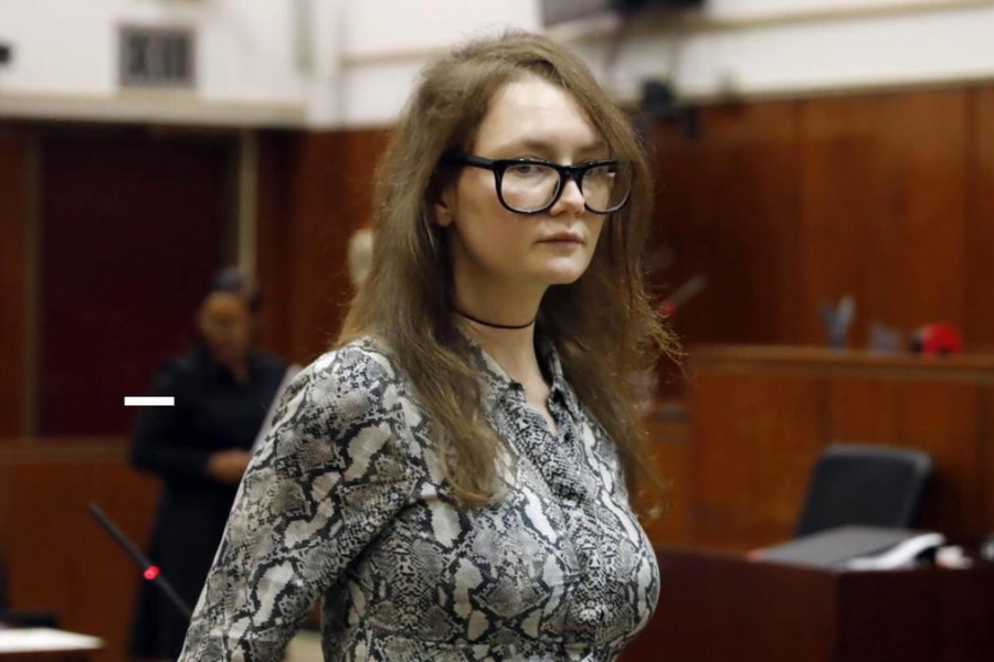 A woman with long, tousled brown hair and black-rimmed glasses stands in a courtroom wearing a fitted snakeskin-print dress and a thin black choker. Her expression is neutral as she looks ahead, with wood-paneled walls and a blurred figure in the background suggesting a legal proceeding.