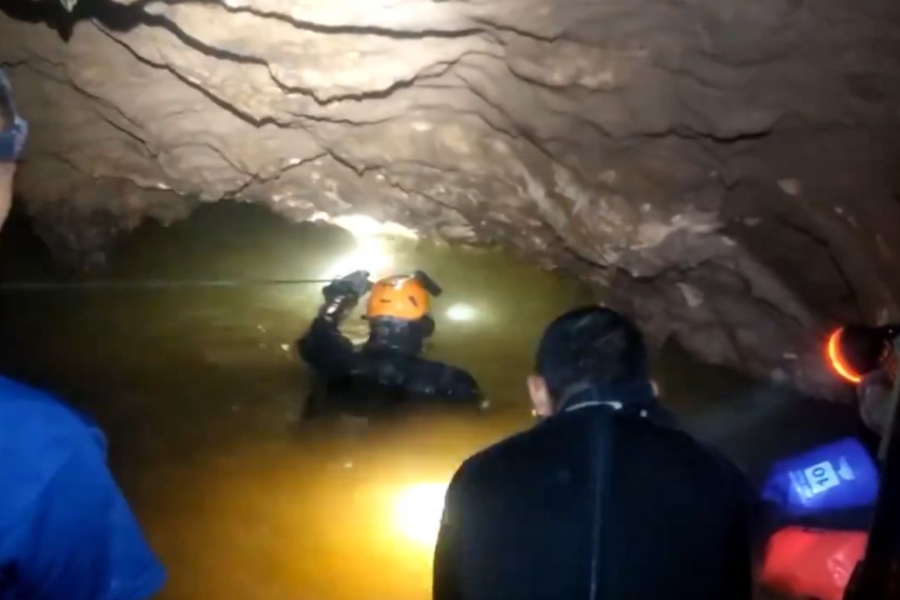 Rescuers equipped with headlamps and wetsuits prepare to navigate a narrow, flooded cave passage. One diver in an orange helmet leads the way into the water, holding onto a guide rope, while others watch or follow closely behind in the dimly lit tunnel.