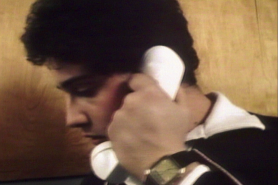 Young man in a collared shirt holds a white corded telephone to his ear while looking down pensively, with a gold digital wristwatch visible on his wrist. The background features a plain wooden wall.