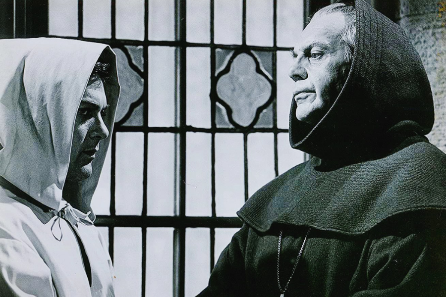 Black and white film still showing two men in hooded monastic robes standing in front of a stained glass window. One wears a light-colored hood, looking intently at the older man in a dark hooded robe, who appears to be in a position of authority.