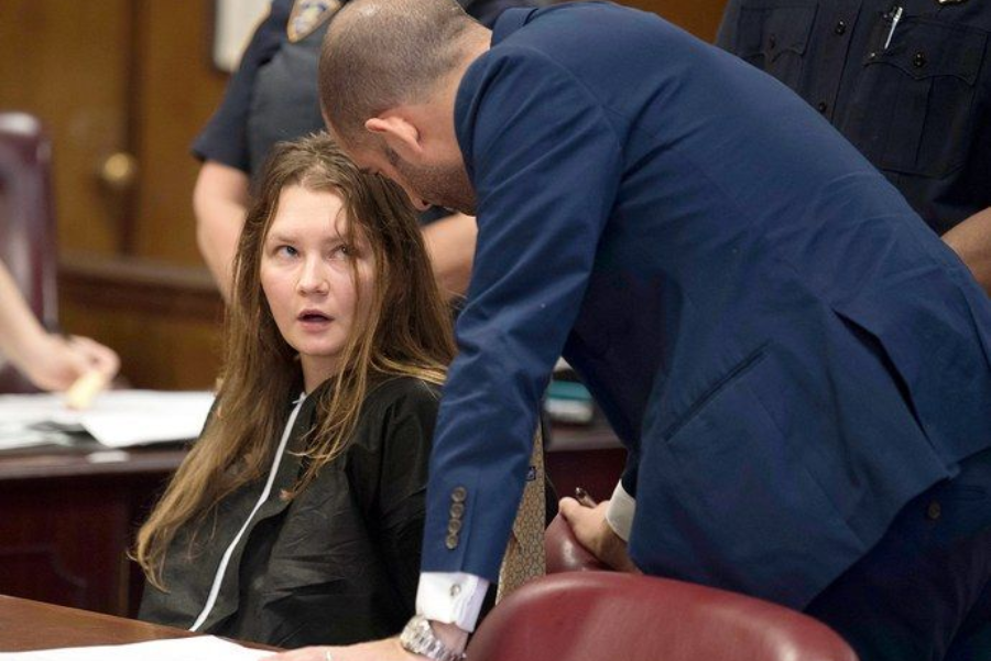 A woman in a black prison-issued outfit sits in a courtroom, looking up with a surprised expression as a man in a blue suit, likely her lawyer, leans over to speak with her. Papers and legal documents are spread out on the table, and uniformed officers stand in the background.