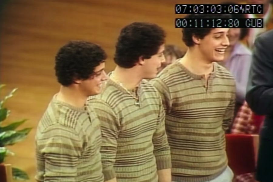 Three young men with similar features and curly dark hair stand side by side, all smiling and wearing matching striped sweaters. The setting appears to be a televised event or show, with timestamp overlays visible at the top of the image.