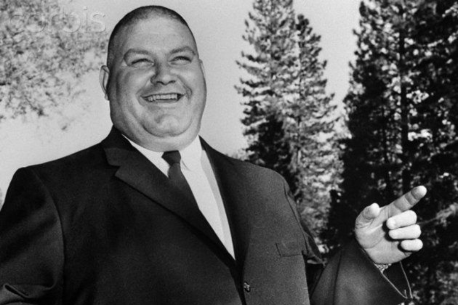 Black and white photo of a cheerful bald man in a suit and tie, smiling broadly while pointing off to the side. He’s standing outdoors with tall pine trees in the background.