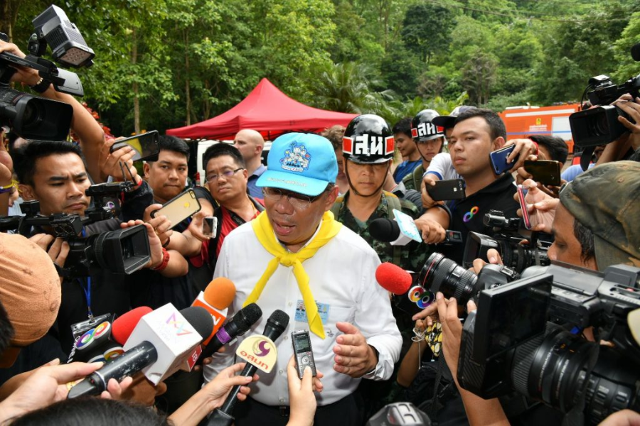 A government official in a blue cap and yellow scarf speaks to a large crowd of reporters and cameramen during a press briefing in a forested area. Microphones and cameras from various media outlets surround him as he addresses the public about the ongoing cave rescue operation.