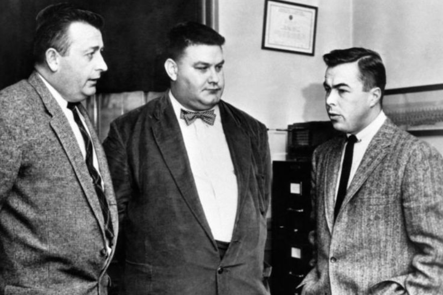 Black and white photo of three men in suits and ties standing indoors, engaged in serious conversation. The man in the center, wearing a bow tie and corduroy jacket, appears to be the focus of the interaction, while office furnishings and a framed certificate are visible in the background.