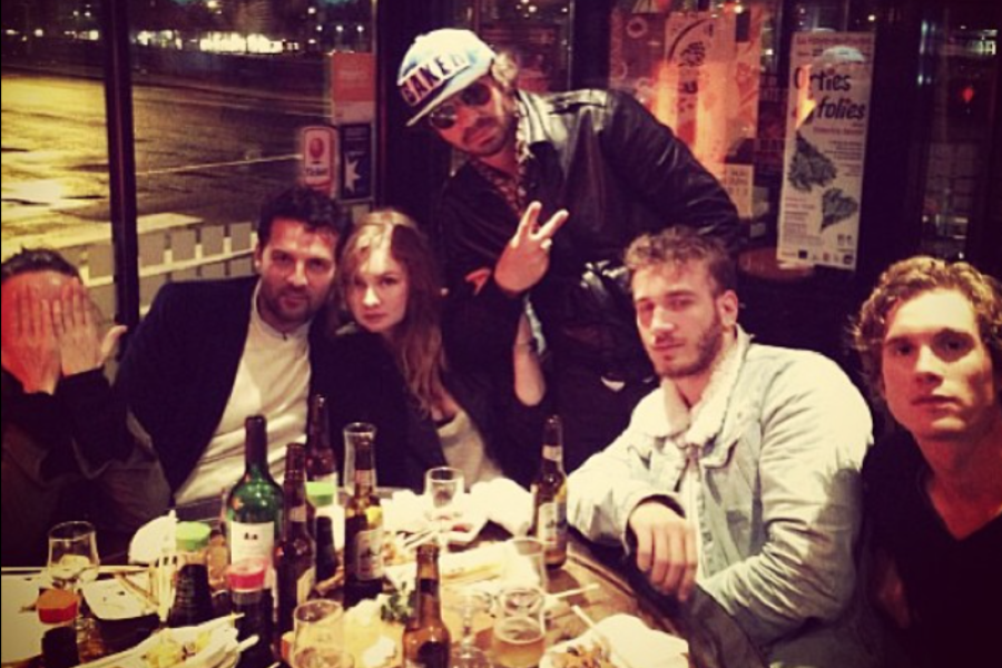 A group of six friends pose around a table cluttered with bottles, glasses, and food at a dimly lit restaurant or bar. One person covers their face with their hands, while another stands and flashes a peace sign wearing sunglasses and a cap that says “BAKED.” The mood appears lively and informal.