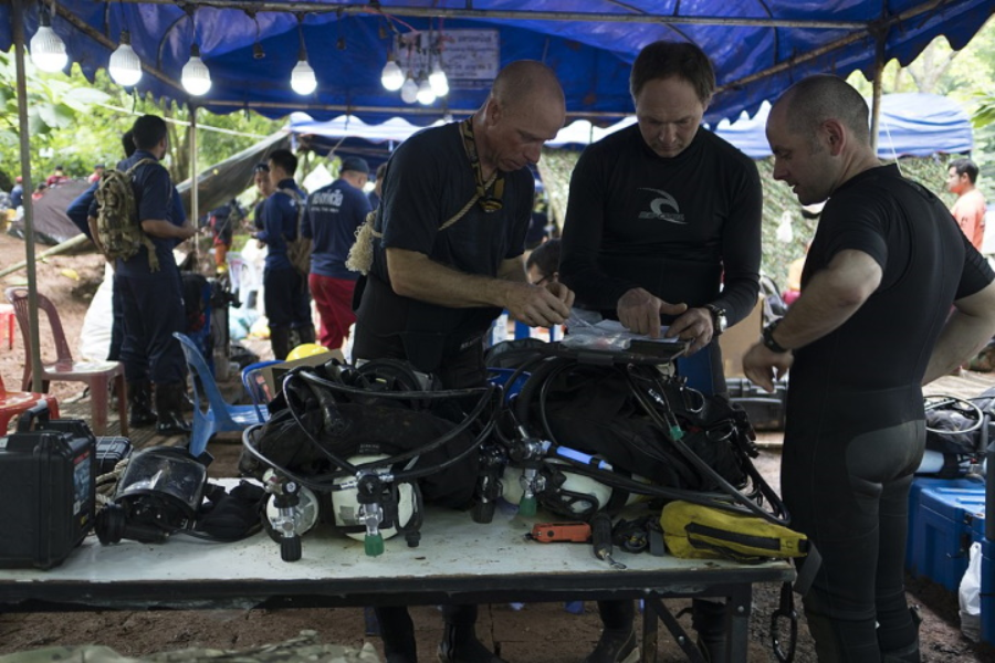 Three rescue divers in wetsuits gather under a blue canopy tent, checking and preparing scuba gear on a table filled with oxygen tanks, masks, and other diving equipment. The background shows a busy staging area with other personnel and gear amid a forested rescue base.