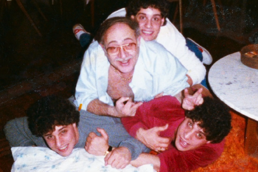 Four people lie playfully stacked on the floor in a cozy indoor setting—three young men with curly hair surround an older man in glasses and an open shirt, all smiling and giving thumbs up or peace signs.
