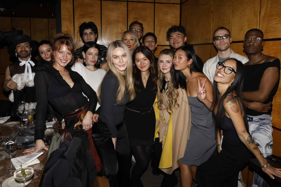 A large group of stylishly dressed young adults pose together inside a warmly lit restaurant, smiling and striking playful poses. The group includes individuals wearing bold outfits and accessories, with one person flashing a peace sign and others leaning in close, creating a lively and celebratory atmosphere.