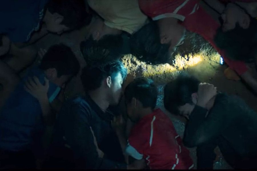 A group of boys and their coach lie huddled together on the muddy floor of a dimly lit cave, conserving warmth and energy. A faint beam from a flashlight illuminates the wet ground near their heads, highlighting the seriousness and vulnerability of their situation.