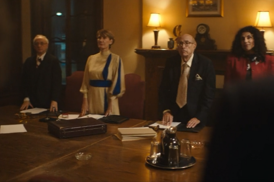 Four formally dressed individuals stand behind a long wooden conference table in a warmly lit room, facing someone out of frame. Papers, folders, and a briefcase lie on the table, suggesting a serious or formal meeting.
