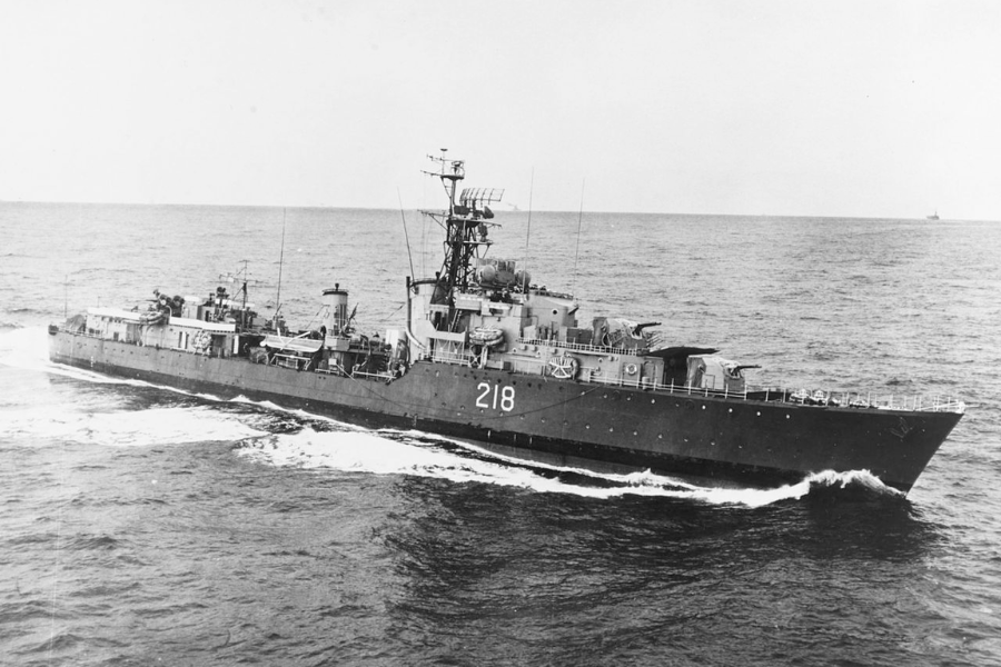 Black and white photo of a naval warship marked with the number “218” cruising through open waters. The ship features multiple antennas, turrets, and lifeboats, with a faint second vessel visible on the distant horizon.