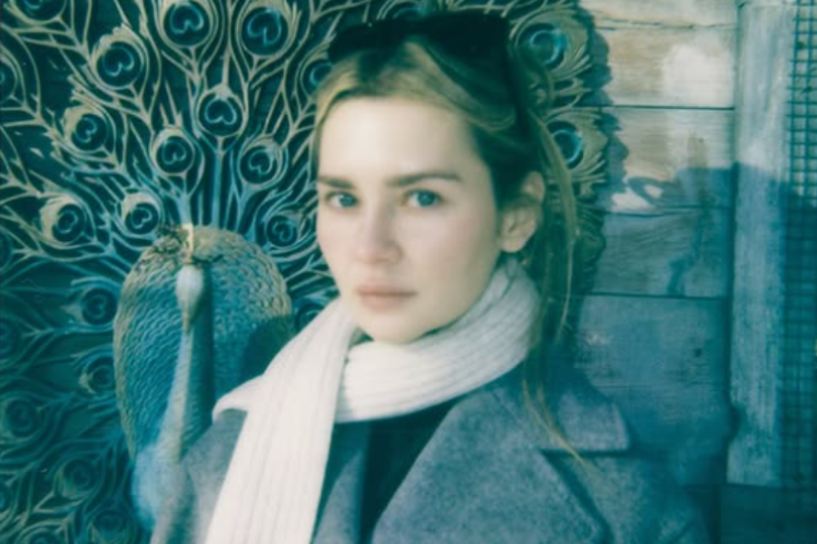 A woman wearing a grey coat and white scarf stands in front of a decorative peacock wall art, with the feathers radiating behind her in a striking pattern. The photo has a soft, vintage filter that adds a dreamy, slightly nostalgic tone.