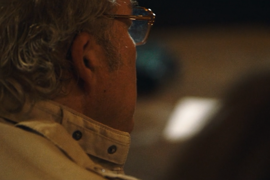 Close-up of an older man seen from behind, wearing glasses and a beige collared shirt with button details. The soft lighting and blurred background suggest an indoor, possibly formal or reflective setting.