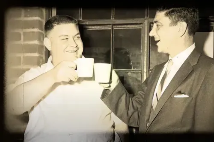 Sepia-toned vintage photo of two men smiling and clinking coffee cups in a cheerful toast. One man wears a short-sleeve shirt, the other a suit and tie, standing in front of a window with brick walls.