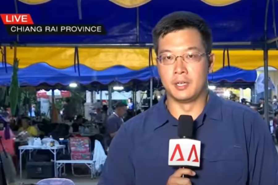 A news reporter stands in front of a busy tented area, holding a microphone with the Channel NewsAsia logo, delivering a live broadcast from Chiang Rai Province. Behind him, rescue personnel and volunteers can be seen working under blue and yellow tents, suggesting an ongoing emergency or rescue operation.
