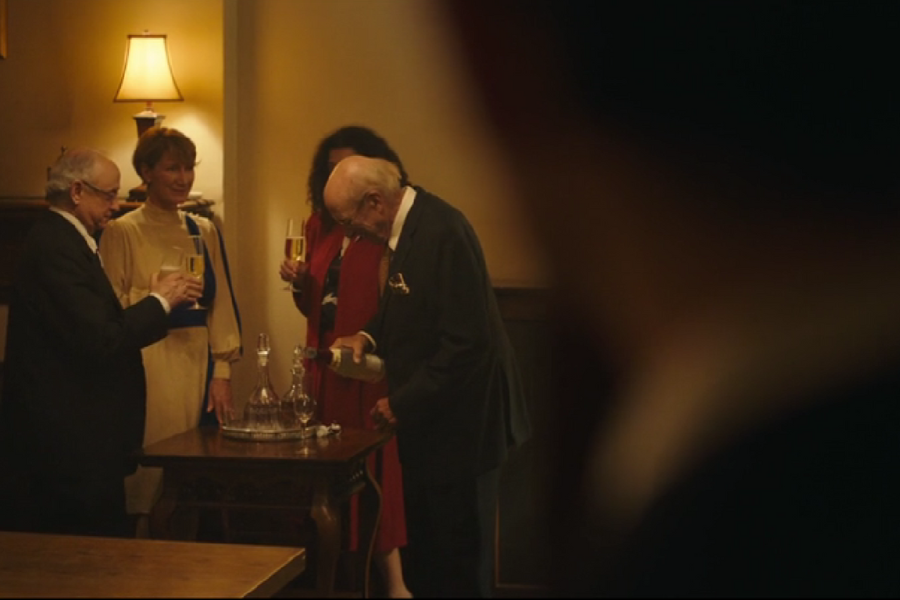 Four well-dressed adults gather around a small wooden table with a decanter set, as one man pours drinks and the others hold champagne flutes. The warmly lit room and elegant attire suggest a celebratory or formal occasion.