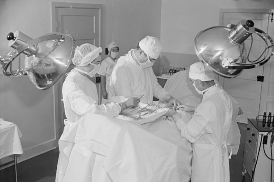 Black and white photo of a surgical team performing an operation in a hospital operating room. Several medical professionals in full scrubs, masks, and caps are gathered around a patient on the operating table under large overhead lights.