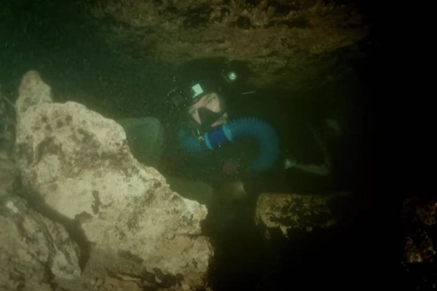 A cave diver wearing a full-face mask and rebreather system maneuvers through a tight underwater passage surrounded by jagged rocks. The diver’s flashlight illuminates the narrow, murky space, emphasizing the challenging conditions of the submerged cave environment.