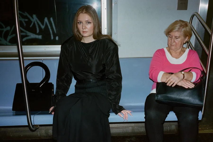 A young woman in a sleek all-black outfit sits on a subway bench next to an older woman in a pink and white top, who is looking down with her arms crossed over her handbag. The young woman faces forward with a composed expression, and a designer bag with a circular handle rests beside her against the subway pole.
