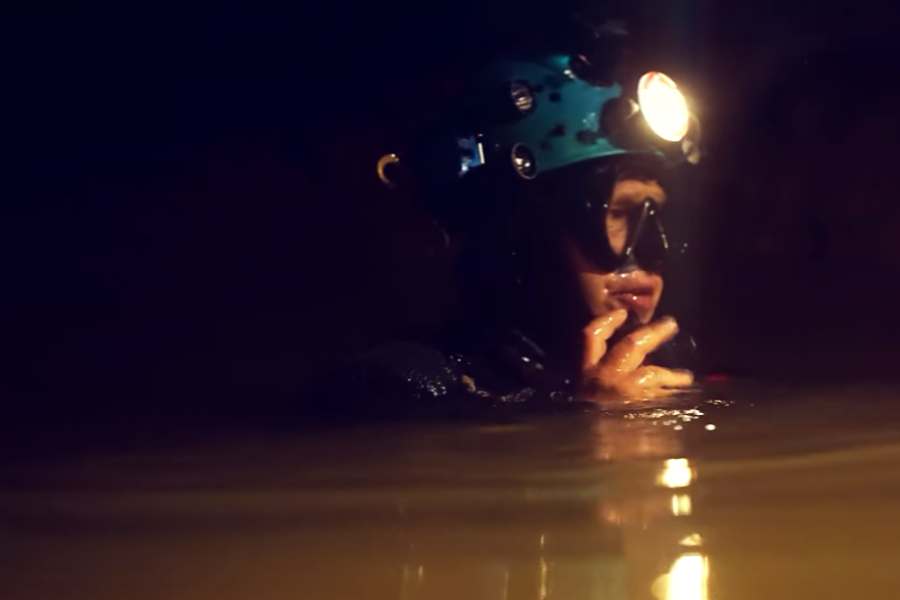 A cave diver wearing a teal helmet with mounted lights and a black diving mask prepares for a dive in dark, murky water inside a cave. The diver's face is partially submerged, with one hand adjusting the mask or regulator, illuminated by the helmet light in the dim environment.