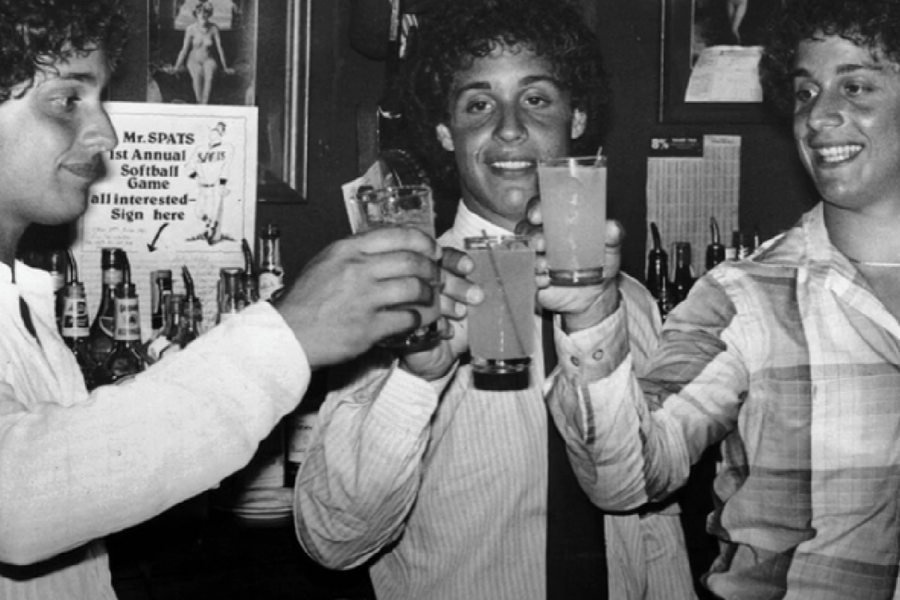 Black-and-white photo of three young men with curly hair toasting with cocktails at a bar, all smiling and dressed in collared shirts. Behind them, a sign reads “Mr. SPATS 1st Annual Softball Game – all interested – Sign here.”
