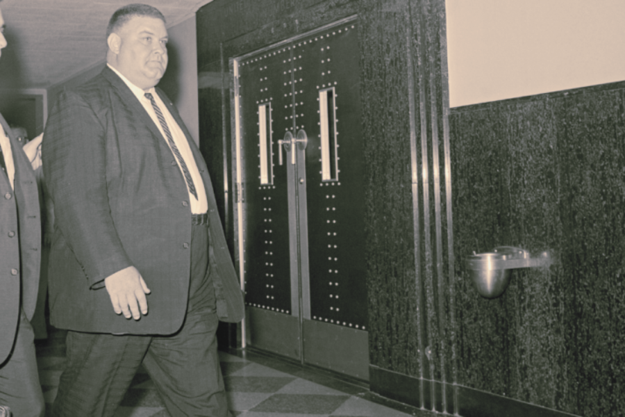 Vintage color-toned photo of a large man in a dark suit and striped tie walking through a hallway with another suited individual partially visible beside him. They pass by large metal double doors and a wall-mounted drinking fountain.