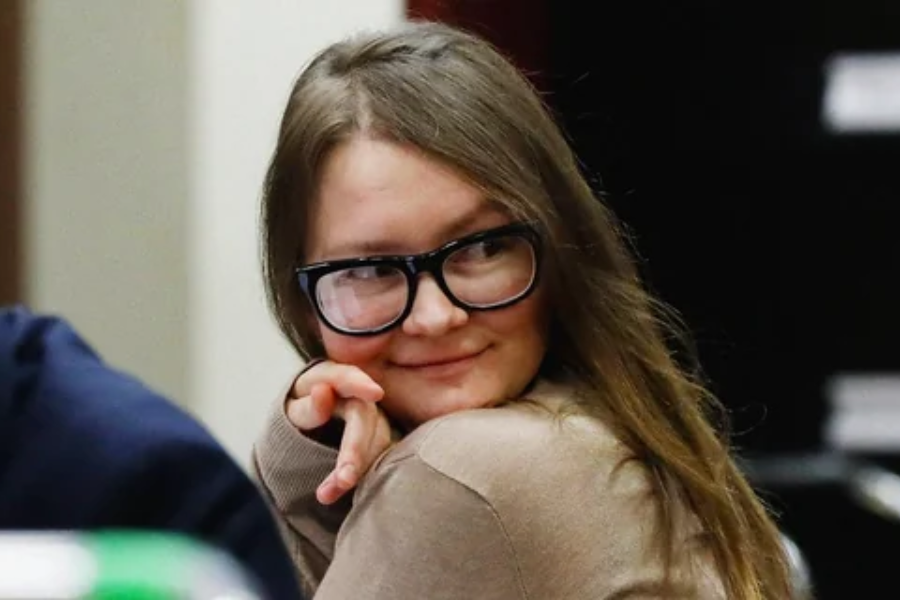 A young woman with long hair and oversized black glasses smiles slyly while glancing over her shoulder in a courtroom. She rests her chin on her hand and wears a beige sweater, exuding a calm and confident demeanor.