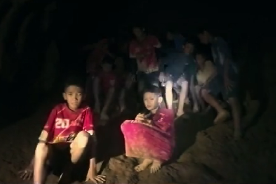 A group of young boys sit and stand on a muddy slope inside a dark cave, illuminated by a flashlight. Some wear soccer jerseys, and all appear barefoot and weary, suggesting they’ve been trapped for an extended period and are awaiting rescue.