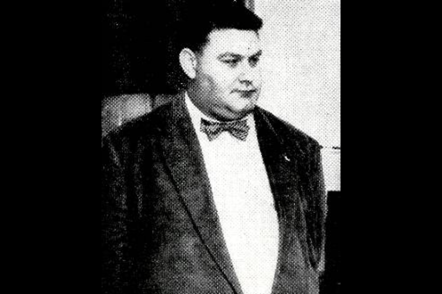Grainy black and white photo of a man in a corduroy jacket and bow tie, standing with a neutral expression. The background appears to be indoors, possibly an office or institutional setting.