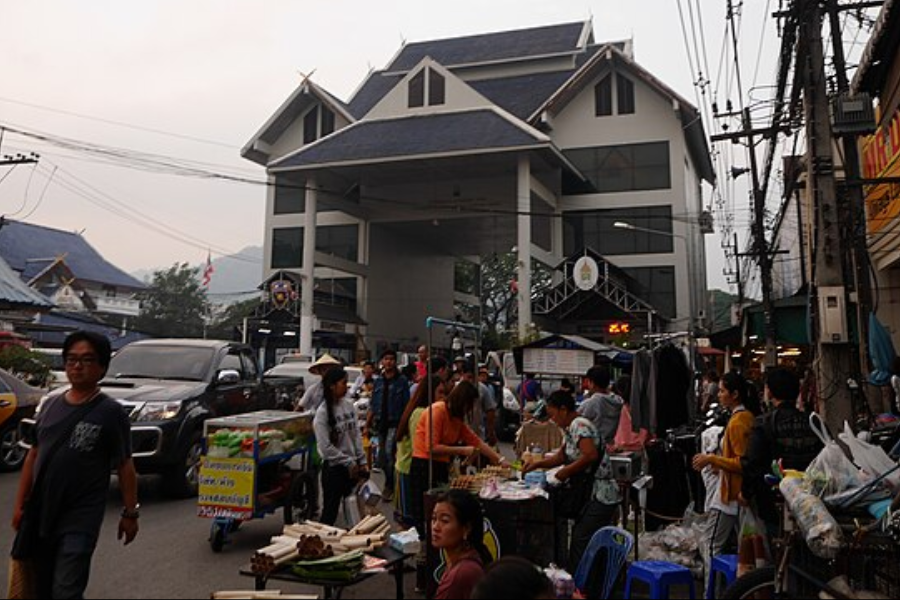 A bustling street market scene in front of a large, traditional-style building with a blue-tiled roof, featuring numerous vendors selling food and goods under makeshift stalls. People walk and shop among tightly packed stalls, with signs in Thai and utility wires crisscrossing above.