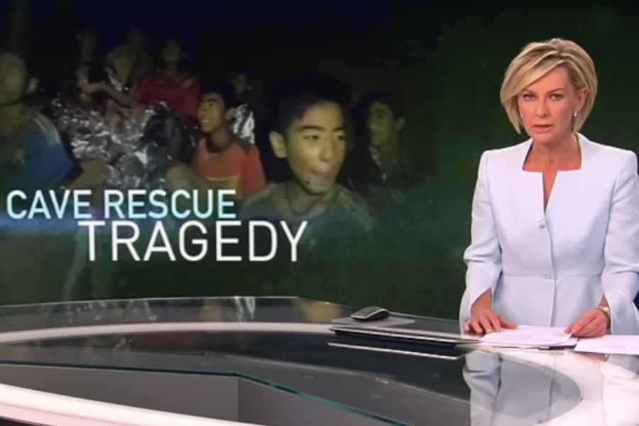 A news anchor in a light blue suit delivers a broadcast titled “Cave Rescue Tragedy,” with an image of rescued boys wrapped in emergency blankets displayed behind her on a large screen. The somber tone and the word “tragedy” imply the report covers a serious incident or loss during the rescue mission.