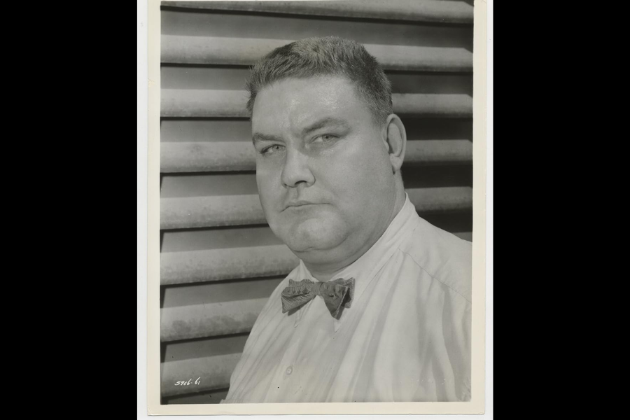 Black and white portrait of a stern-looking man in a collared shirt and bow tie, standing in front of a backdrop of horizontal metal slats. His expression is serious, and the image has a studio-like quality.
