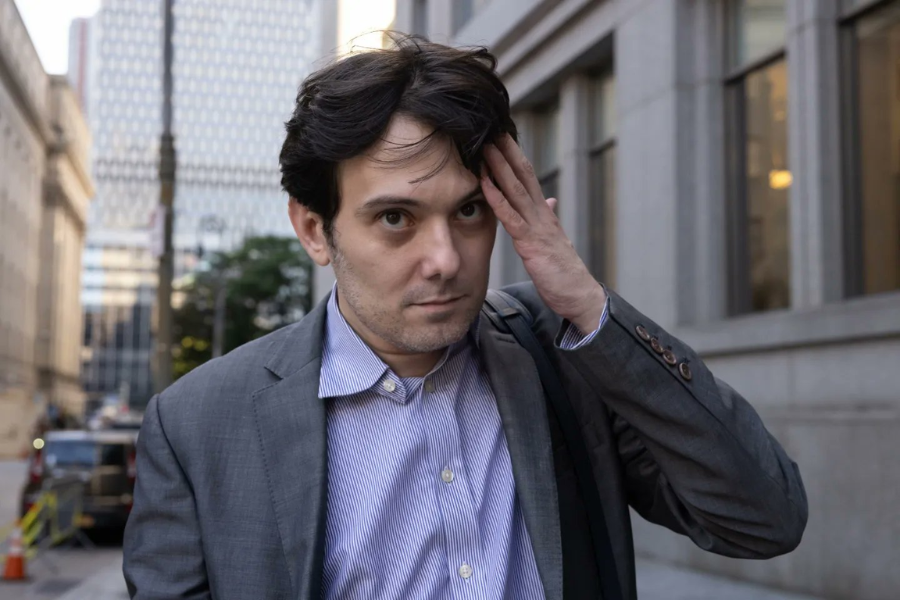 A man in a gray suit jacket and light blue button-up shirt stands on a city sidewalk, brushing his hair back with one hand while looking ahead with a serious expression. Tall office buildings and a few vehicles are visible in the background, indicating an urban downtown setting.