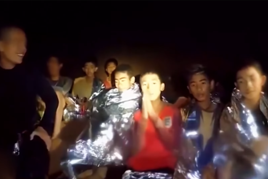 A group of boys wrapped in reflective emergency blankets sit together inside a dark cave, illuminated by flashlight. One boy in a red shirt clasps his hands in a gesture of prayer or gratitude, while others appear calm or relieved, suggesting they’ve just been found by rescuers.
