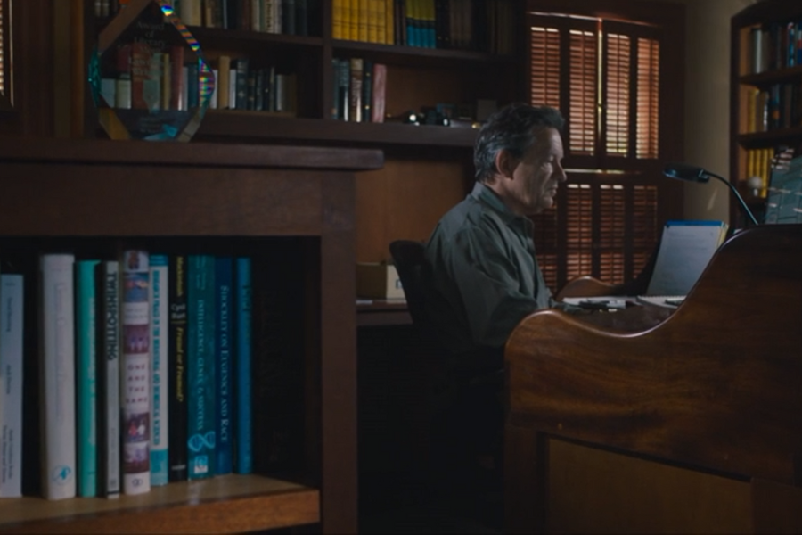 A man sits at a polished wooden desk in a study or office, focused on his laptop with shelves of books and soft window light behind him. A nearby bookshelf prominently displays psychology and social science books, suggesting academic or research work.