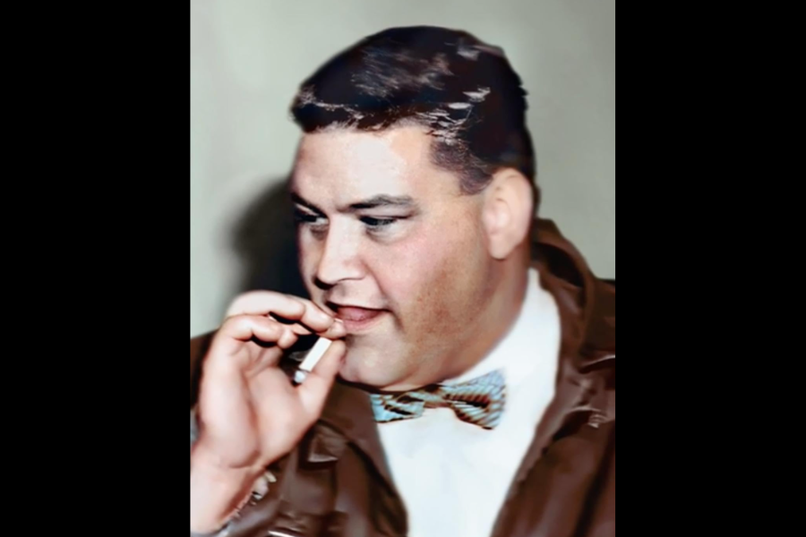 Colorized portrait of a man in a brown jacket and multicolored bow tie, holding a cigarette near his mouth. He appears mid-conversation or thought, looking slightly to the side against a neutral background.