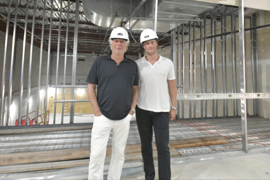 Two men wearing white construction helmets pose inside a building under construction, surrounded by exposed metal framing and ductwork. One wears a black polo shirt with white pants, while the other wears a white polo shirt with black pants; both smile slightly while standing on a partially completed floor.