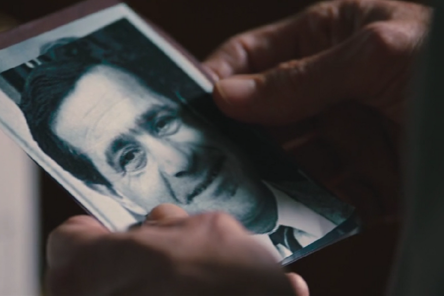 Close-up of hands holding an old black-and-white photograph of a middle-aged man in a suit, with a slight smile and kind expression. The photo appears worn, suggesting it holds sentimental or investigative significance.