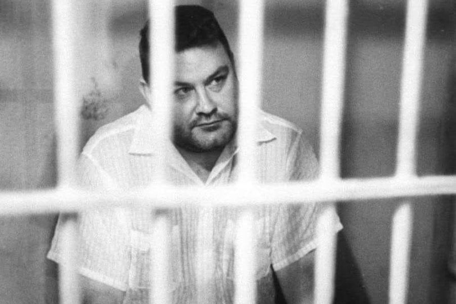 Black and white photo of a man in a short-sleeve button-up shirt sitting behind prison bars. He has a contemplative expression and is partially obscured by the vertical bars in the foreground.