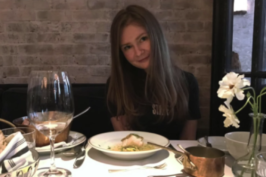 A woman with long straight hair sits at a cozy restaurant table, smiling softly with her head slightly tilted. In front of her is a partially eaten plated dish, a wine glass, a small copper pot, and a white flower in a vase, set against an exposed brick wall.