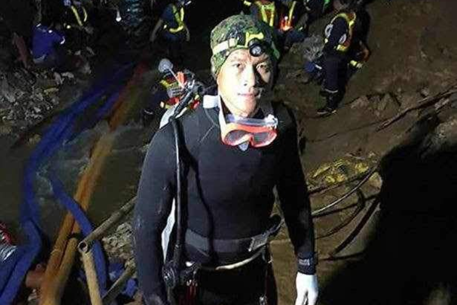 A diver in a black wetsuit with a camouflage beanie, headlamp, and pink goggles around his neck stands in a dimly lit cave. Behind him, rescue team members work near water hoses and muddy terrain, indicating a high-stakes cave rescue operation.