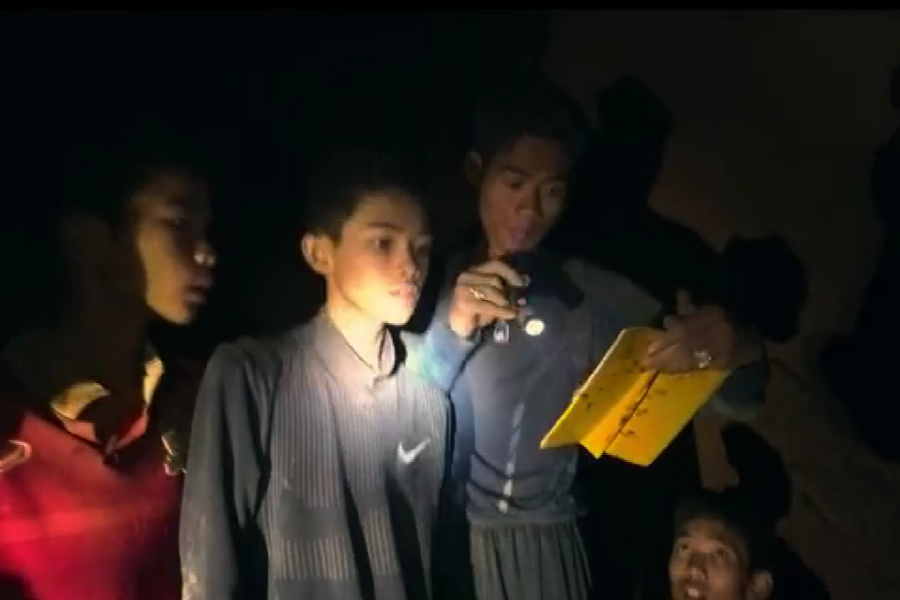 Four boys inside a cave are illuminated by a flashlight, with one boy holding and reading from a yellow notebook. The scene conveys a moment of communication or message delivery during their time trapped underground, reflecting their composure and cooperation.