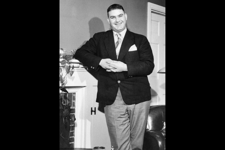 Black and white photo of a tall man in a blazer, tie, and slacks smiling and posing with his hands folded in front of a fireplace mantel. He stands confidently in a cozy, well-furnished room with a visible door and a leafy plant on the mantle.