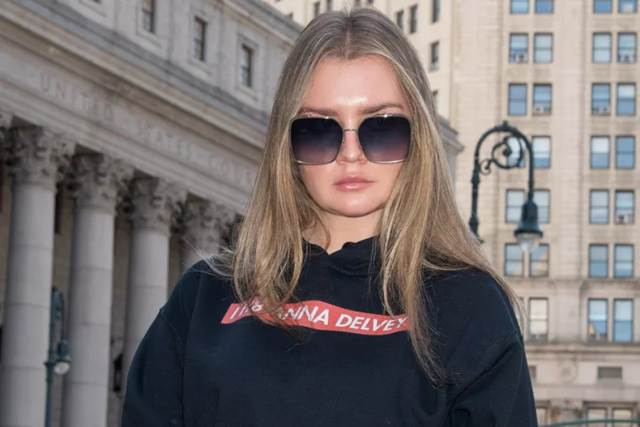 A woman with straight blonde hair wears oversized square sunglasses and a black hoodie with a red label that reads "ANNA DELVEY" in bold white letters. She stands confidently in front of a courthouse, with the engraved words “UNITED STATES COURT” partially visible on the stone building behind her.