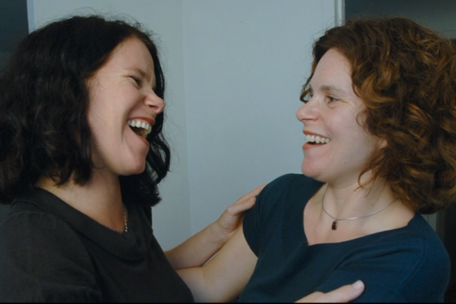 Two women with curly hair smile and laugh while holding each other’s arms, sharing a joyful moment in a brightly lit room. Their expressions convey a sense of warmth, connection, and familiarity.
