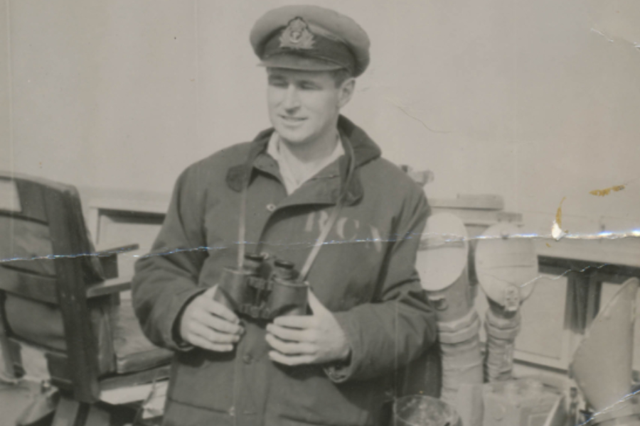 Faded black and white photo of a young man in a naval uniform and cap, holding binoculars on a ship’s deck. He wears a coat marked “RCN” and stands beside nautical equipment, smiling slightly.